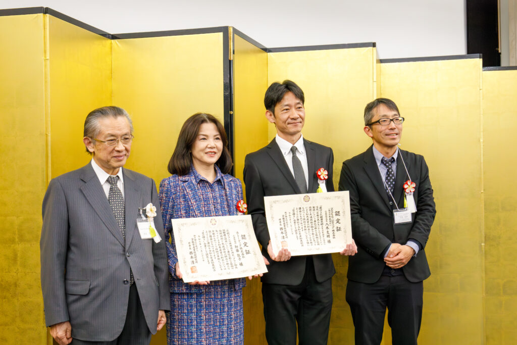 金屏風の前で、認定証を手にした男女2名を含む、スーツやジャケット姿の男女4名が並んで立つ記念撮影の様子。