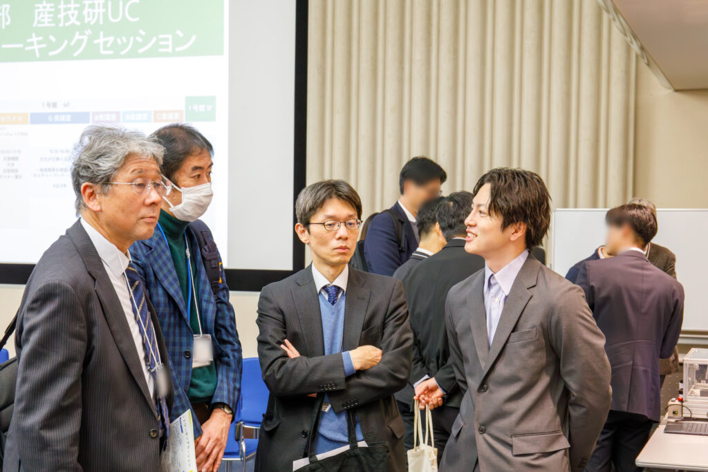 「ワーキングセッション」と映し出されたスクリーンを背景に、スーツ姿の男性数名が集まって真剣な表情で意見交換を行う様子。