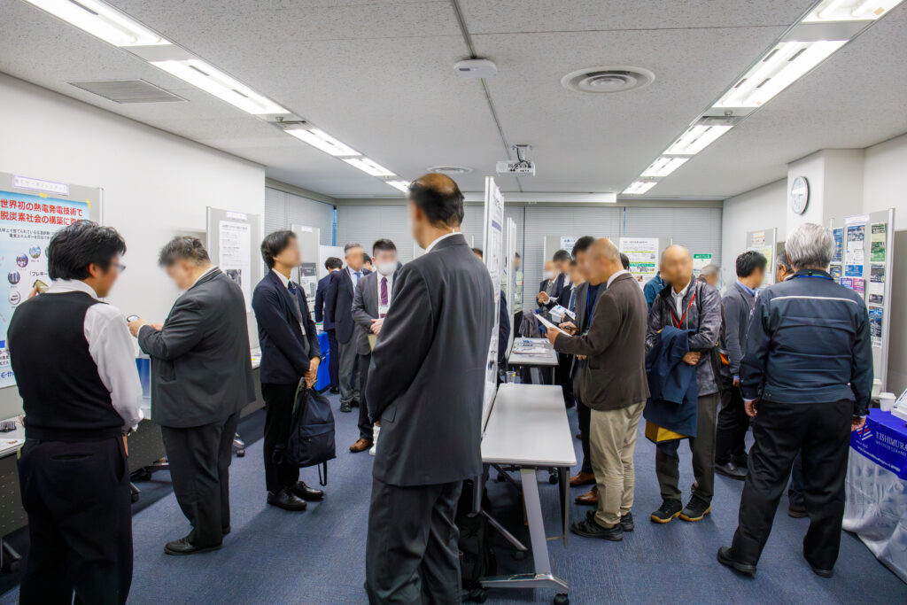 多くの来場者が行き交う展示会場で、壁際や中央に設置されたパネル展示を熱心に見学し、担当者と対話するスーツ姿の人々の様子。