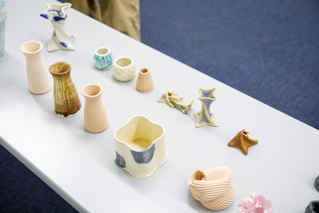白い長机の上に並べられた、3Dプリンターで制作されたと思われる複雑な幾何学模様や積層痕を持つ、色とりどりの陶磁器の造形作品群。
