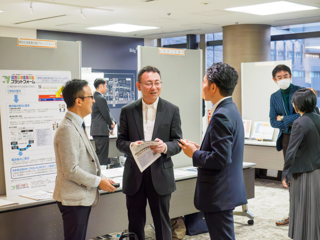 屋内展示会場のブース前で、パンフレットを手に持ち談笑するスーツ姿の男性3名と、周囲で接客や移動を行う参加者の様子。