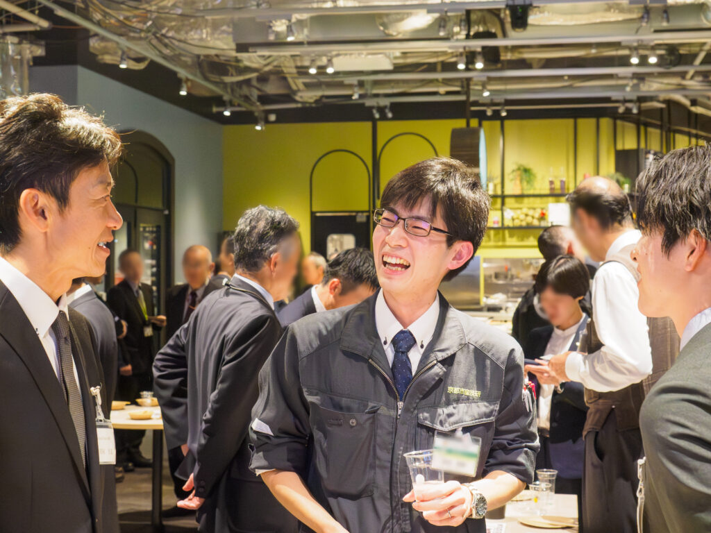 交流会会場にて、「京都市産技研」のロゴ入り作業着を着た男性が、スーツ姿の参加者らと飲み物を手に満面の笑みで談笑している様子。