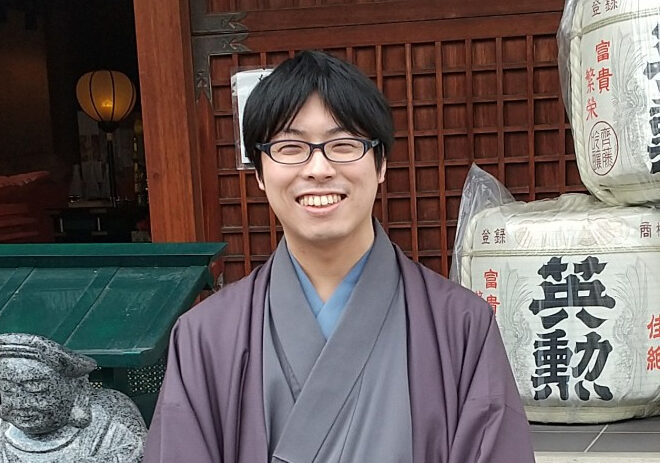 寺社のような建物の前で、着物に羽織をまとった笑顔の男性が、酒樽や大黒天の石像の隣に立っている様子。