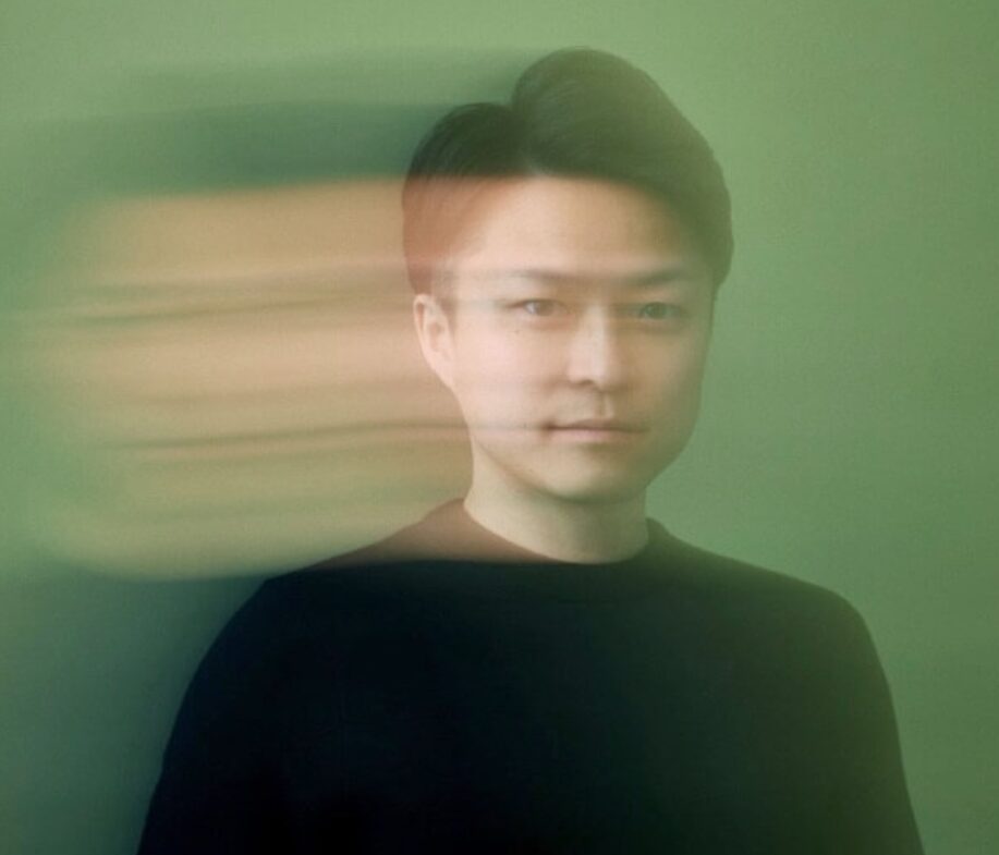 緑色の背景の前で、横にぶれた残像を伴う演出が施された、黒い服を着た若い男性のバストアップ写真。