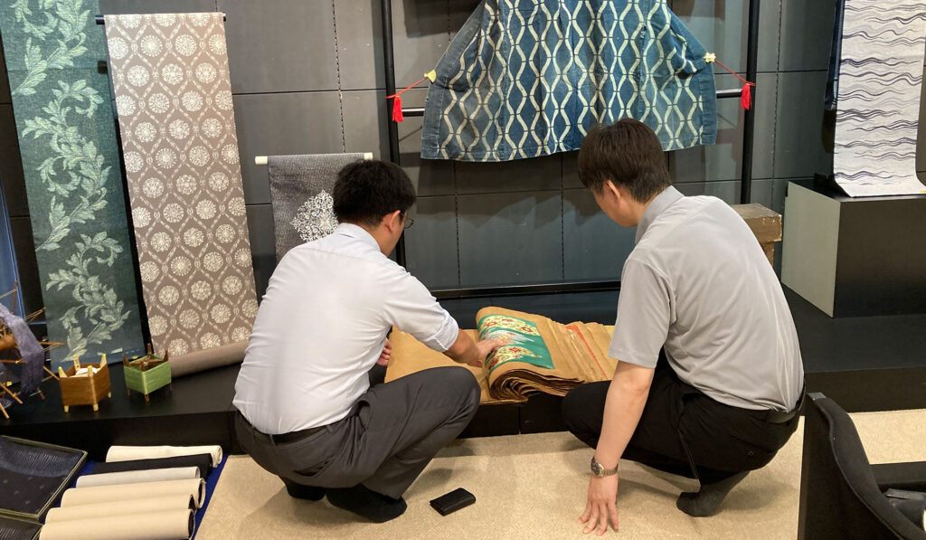 展示スペースにて、伝統的な織物や着物が飾られる中、床に置かれた見本帳を屈んで熱心に確認する2名の男性。