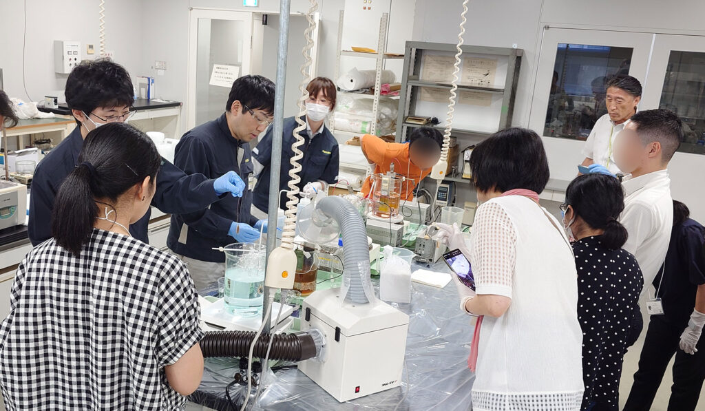 実験室のような施設で、白衣や作業着を着たスタッフがビーカーや装置を使い、見学者に囲まれながら作業や説明を行う様子。